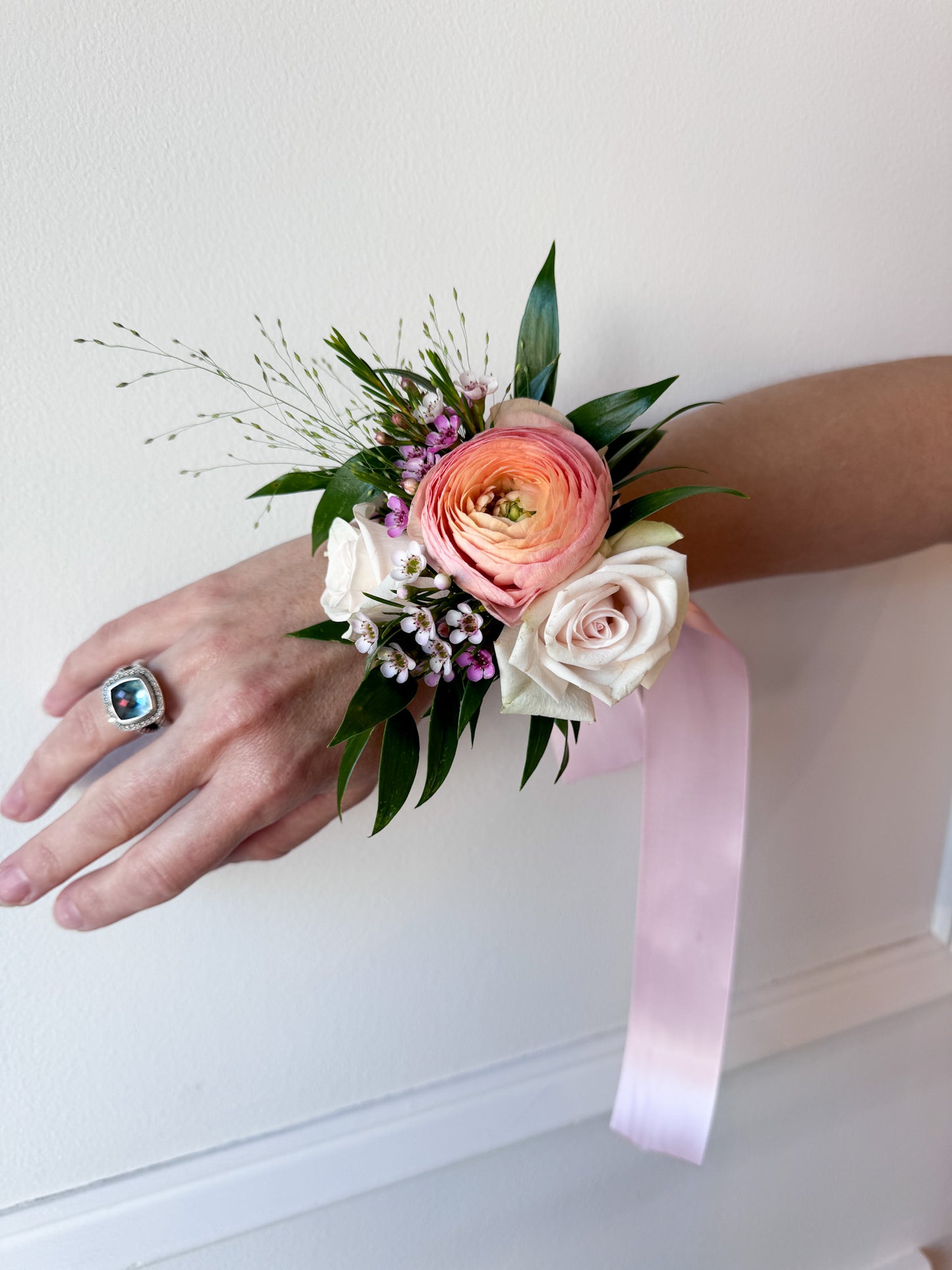 A wrist corsage featuring a mix of pink, white, and peach flowers with greenery, attached to a pink ribbon. Prom, homecoming, wedding corsage