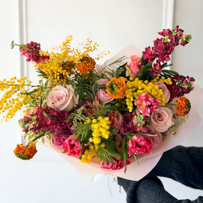 Breathtaking hand-tied garden bouquet featuring yellow Mimosa, pink ranunculus, and purple snapdragons, for March's Monthly Color Palette, wrapped in blush paper for International Women's Day.