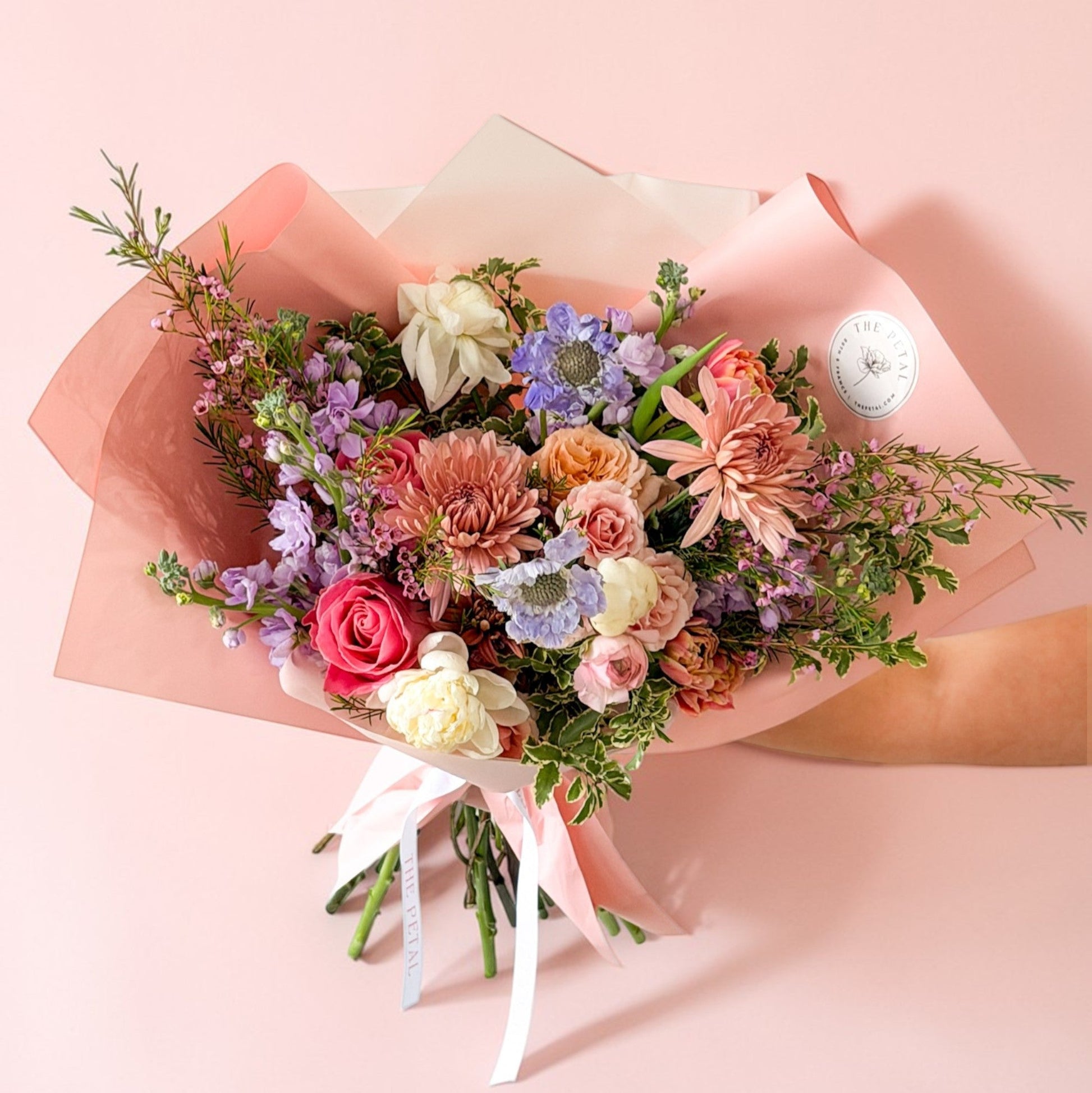 Fresh, organic hand wrapped bouquet of colorful pink, coral, peach, and purple flowers in premium fancy paper for Mother's Day with roses, peonies, ranunculus, and tulips.