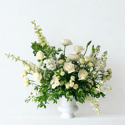 A white floral arrangement with white roses, hydrangeas, and green foliage in a white urn-style vase for sympathy, funeral or celebration of life for delivery in Minneapolis area.