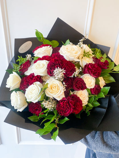 Hand-tied bouquet of white and red roses with accent greenery in black premium wrap, from a women-owned & operated, small business. Shop local and enjoy Valentine’s Day delivery from your trusted Minneapolis florist.