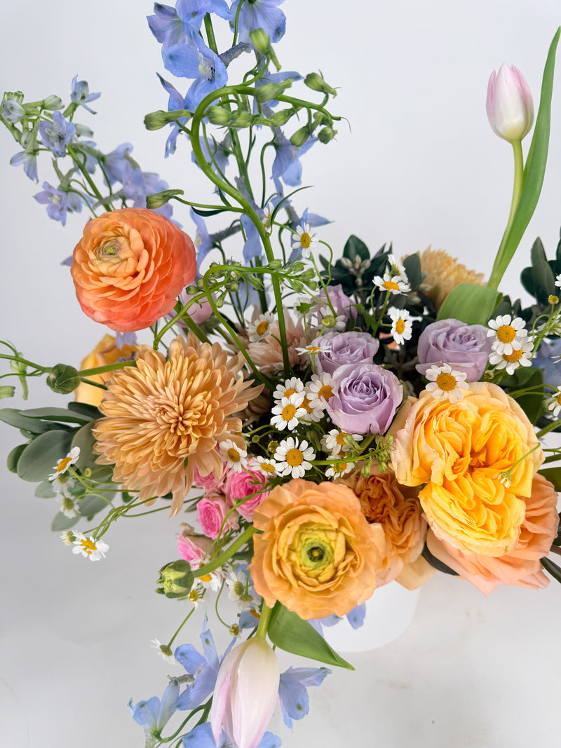 Bouquet of colorful flowers including orange, purple, and yellow on a light background. Ranunculus, tulips, delphinium, feverfew, and roses. spring flowers order for delivery.