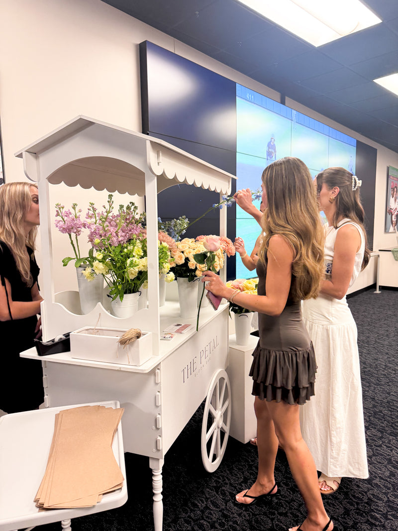 Woman selecting flowers from a white  Bloom Bar flower cart at an event. Bridal shower event idea. Birthday party event idea, baby shower idea.