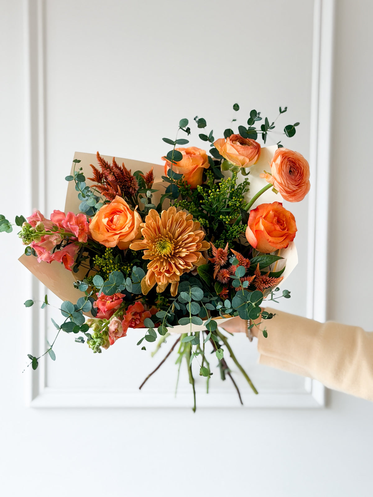 Handcrafted fall bouquet in vibrant coral, mauve, peach, and orange tones with fresh green foliage, wrapped in paper and tied with a ribbon Available for local flower delivery in Minneapolis, Edina, Wayzata, and Minnetonka.