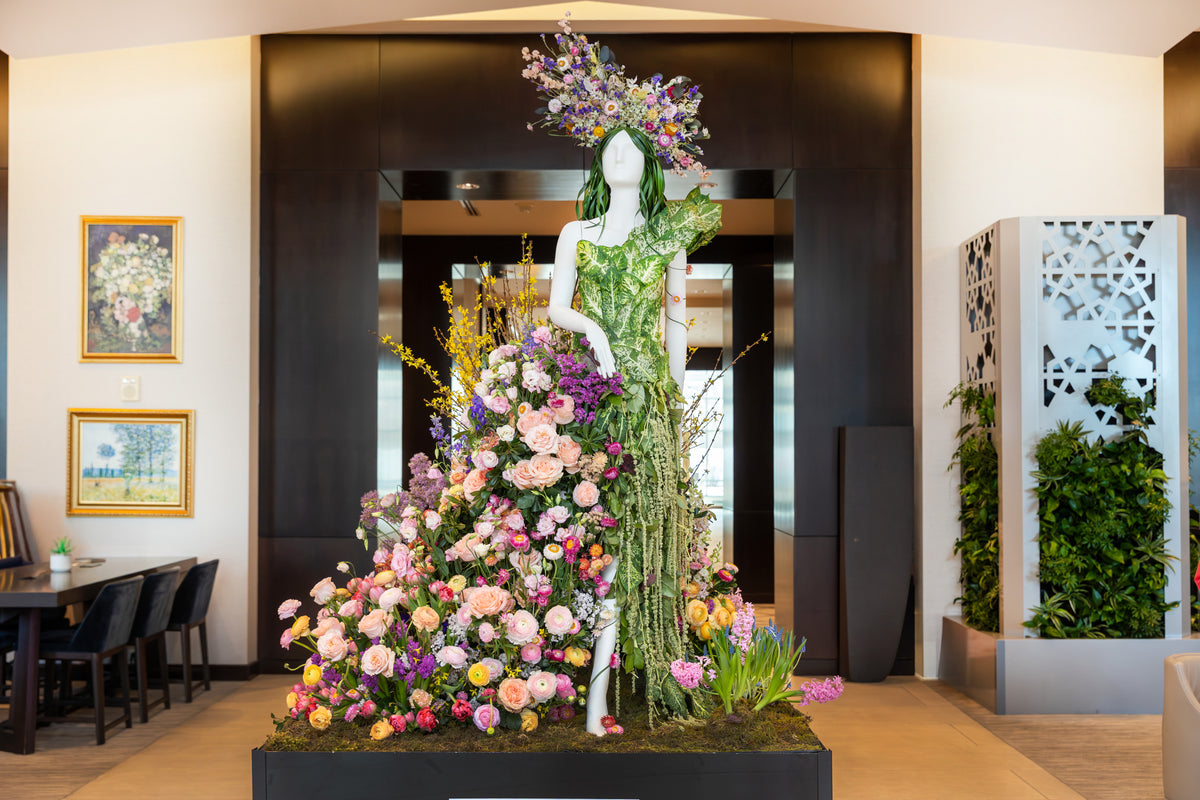 A large floral sculpture of a woman in a green dress stands on a black base in a modern indoor lobby, surrounded by abundant pink, purple, yellow, and white flowers.