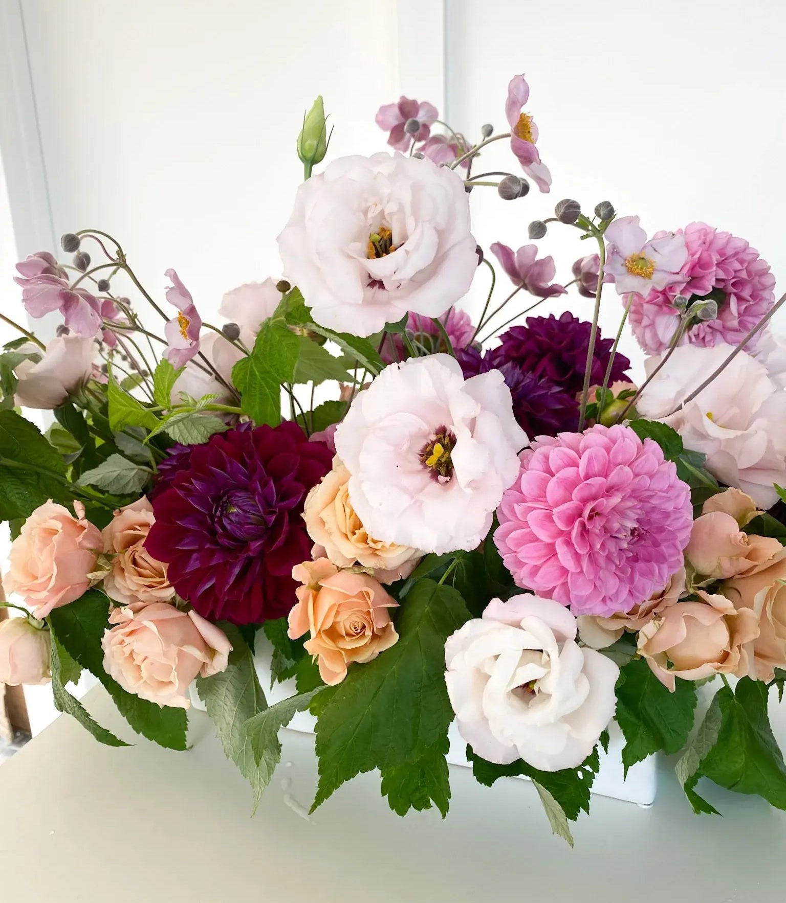 A lush floral centerpiece with burgundy dahlias, peach spray roses, and pale pink lisianthus in a white container. Order Flowers for delivery Twin Cities area. 