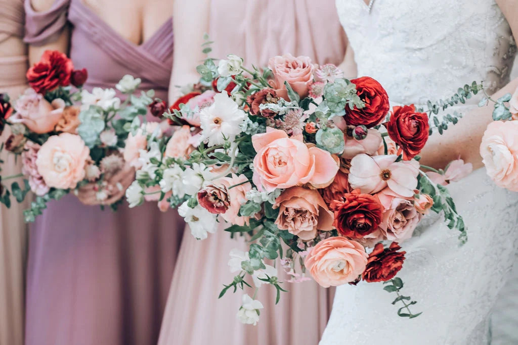 Close-up of a bride and bridesmaids holding textured wedding bouquets with deep red roses, peach ranunculus, and eucalyptus.