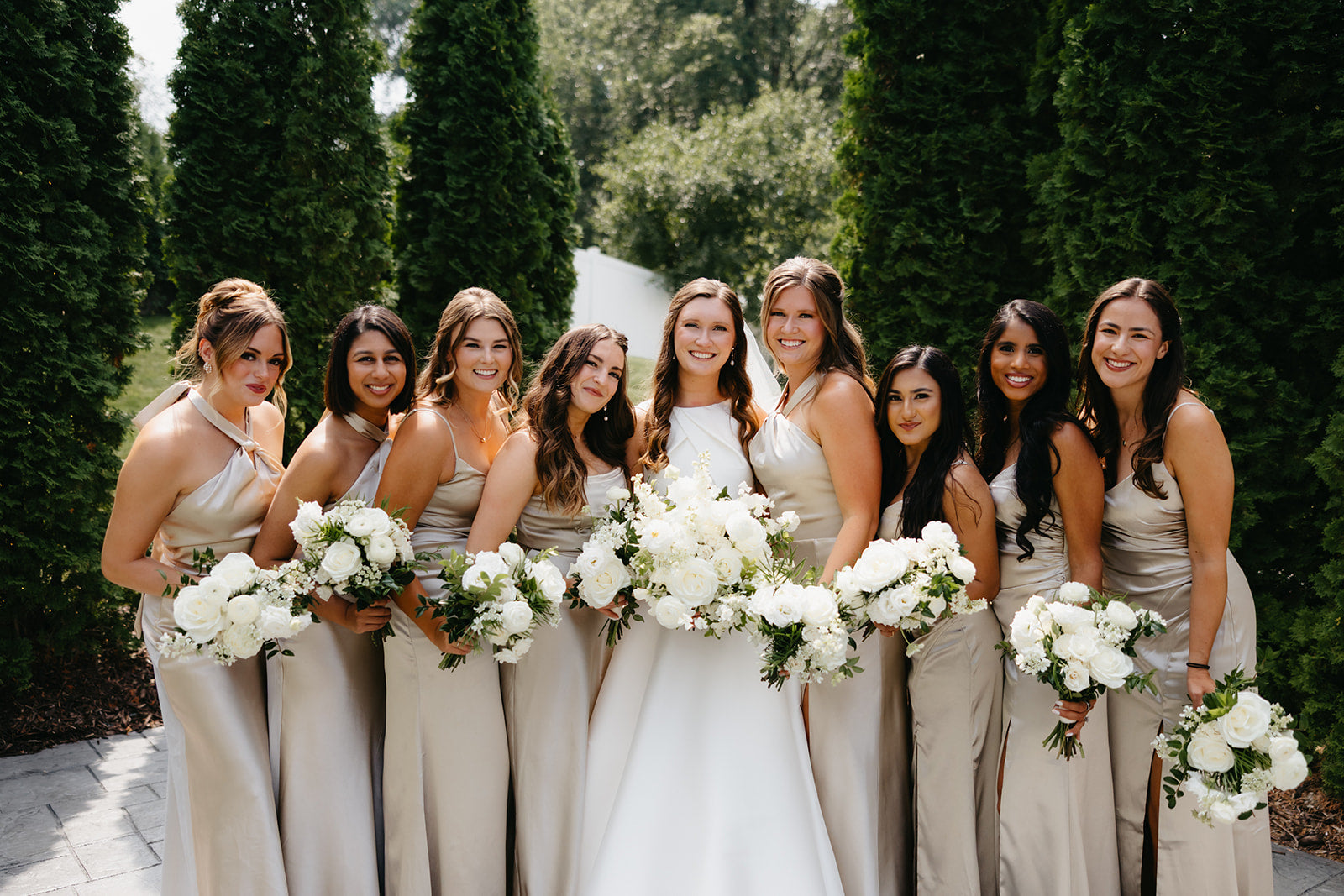 Bride with bridesmaids in champagne dresses holding white bouquets outdoors.