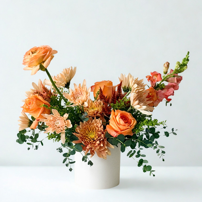 Handcrafted October floral arrangement in vibrant coral, mauve, peach, and soft orange tones with lush green foliage in a modern white vase. Available for local flower delivery in Minneapolis, Edina, Wayzata, and Minnetonka.