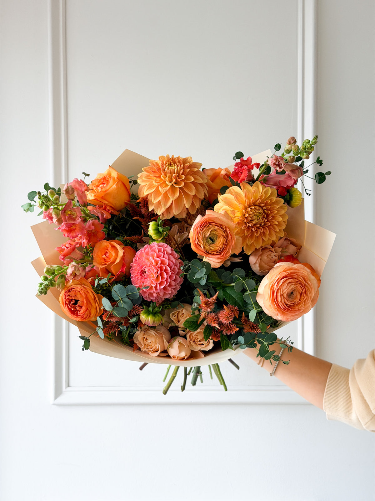 Handheld Deluxe October bouquet featuring coral, mauve, peach, and soft orange hues with fresh green foliage, wrapped in paper and tied with a ribbon. Available for delivery to Hopkins, Minneapolis, Plymouth, and Wayzata.