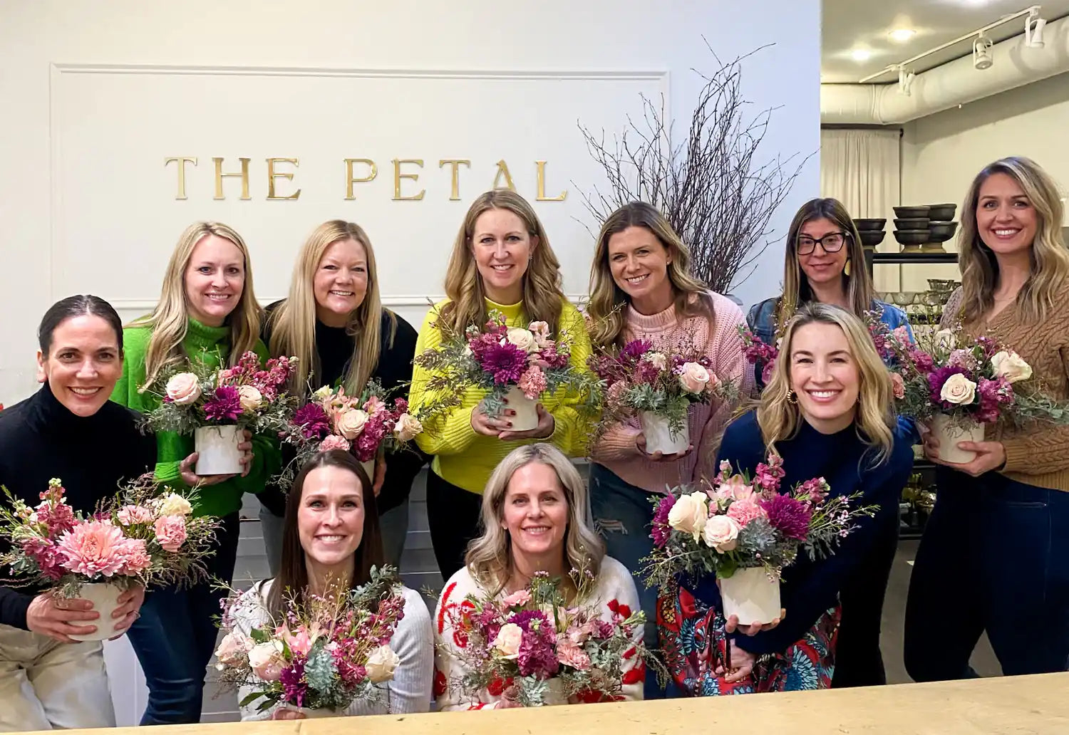 A group of women smiling and holding their finished pink and purple floral arrangements at a design workshop at The Petal.