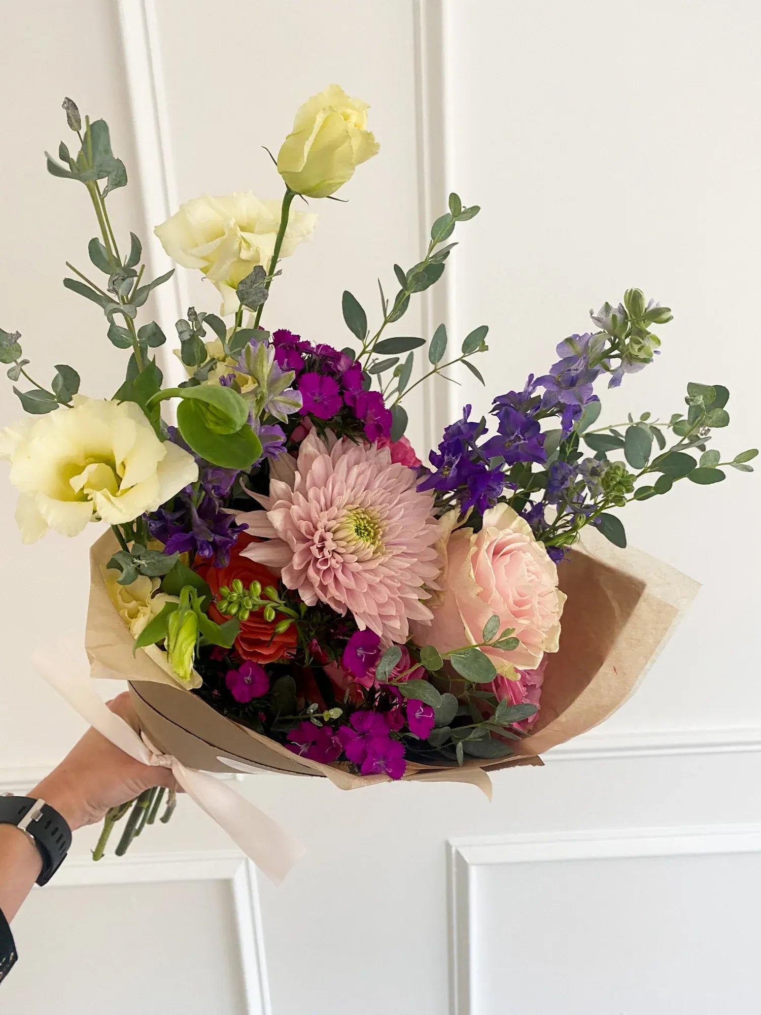 A hand-tied floral bouquet with a large pink chrysanthemum, yellow lisianthus, and purple filler flowers wrapped in brown kraft paper.