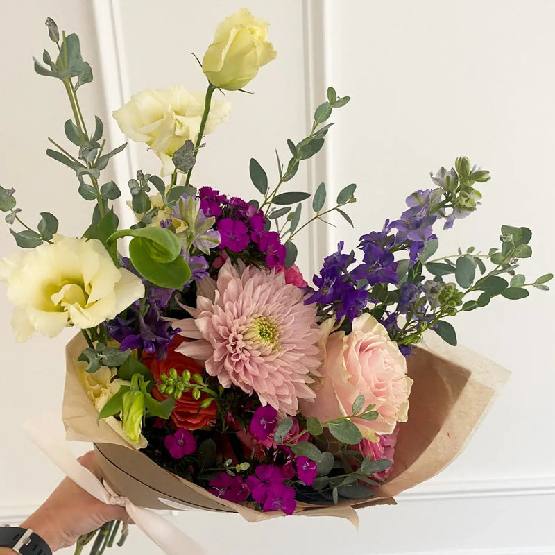 A hand-tied floral bouquet with a large pink chrysanthemum, yellow lisianthus, and purple filler flowers wrapped in brown kraft paper for a bloom bar event in Minnetonka or Edina.