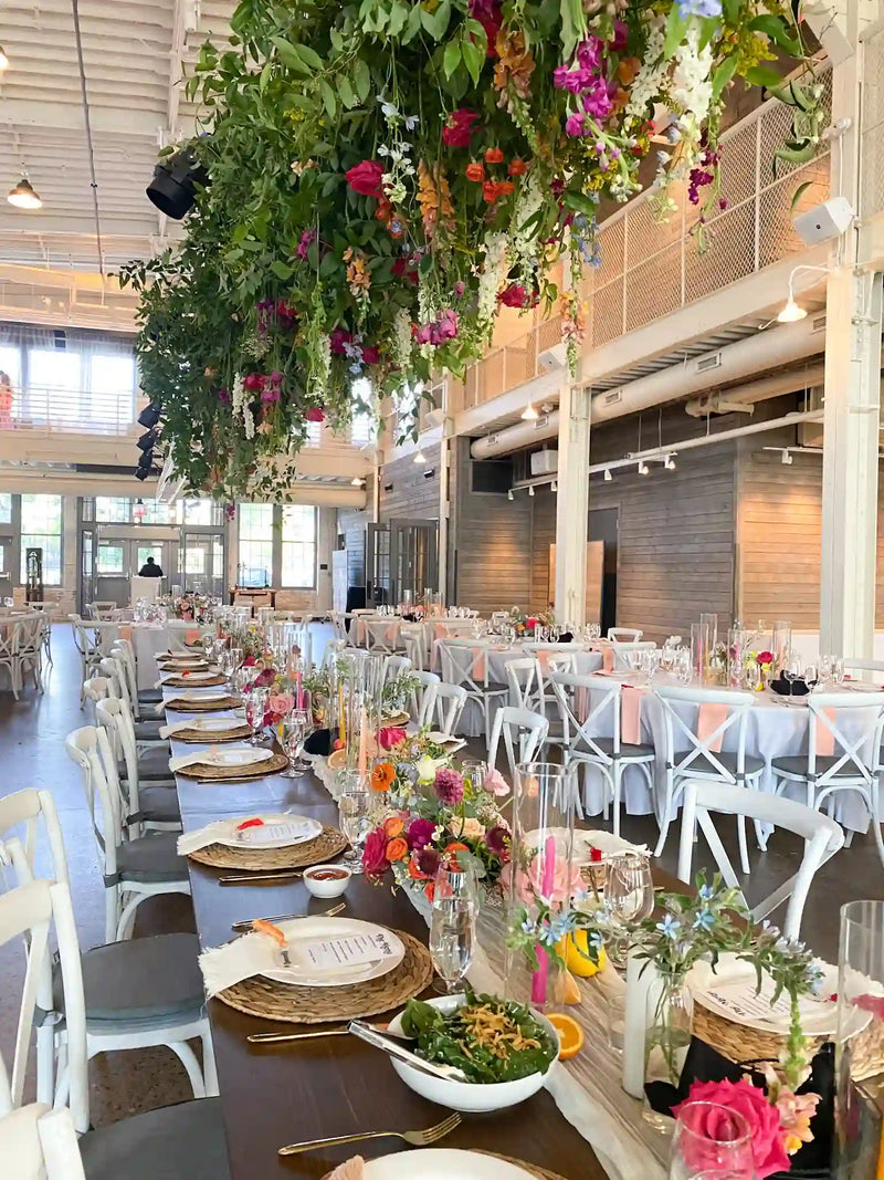 Wide view of an industrial wedding reception space with long wooden tables and a massive overhead hanging greenery and floral installation.