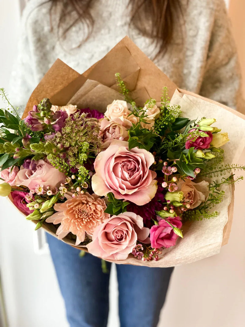 A romantic bouquet featuring mauve roses, peach mums, and delicate green foliage wrapped in rustic brown paper.