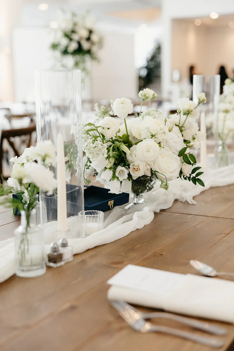 Lush white floral compote centerpieces and bud vase in glass vases on a long wooden reception table with a white fabric runner and taper candles. Hutton House. Head Table Flowers