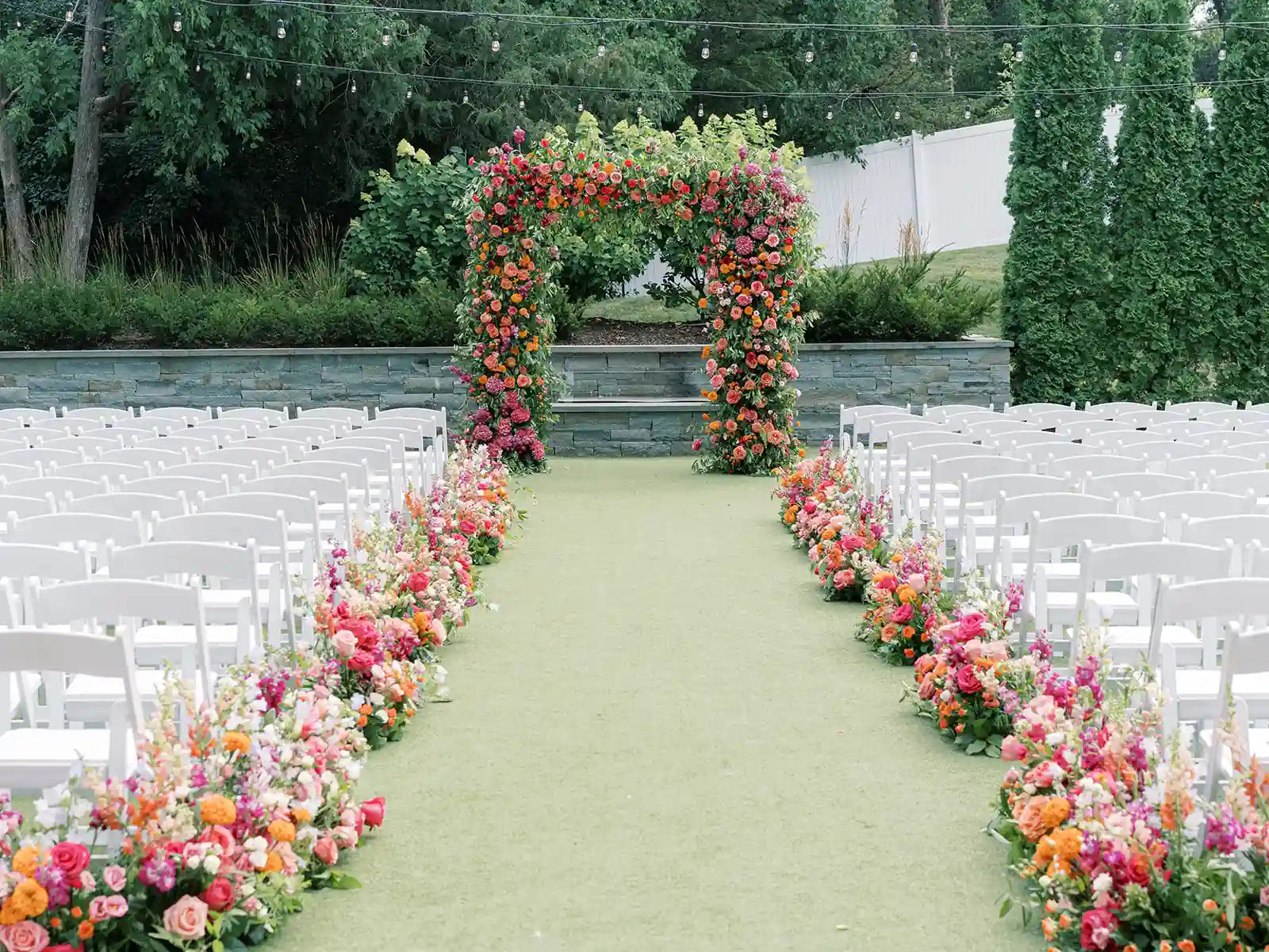 Vibrant outdoor wedding ceremony setup with a full floral arch and matching flower meadows lining a grass aisle with white chairs.