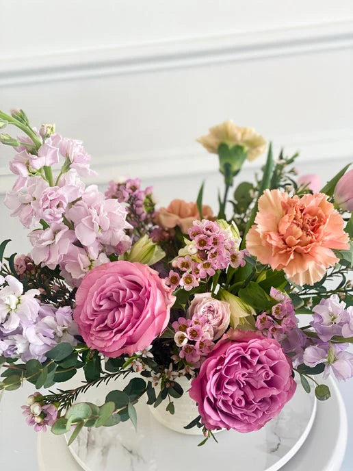 Close-up of a floral arrangement with pink garden roses, peach carnations, and purple stock flowers in a white ceramic vase.