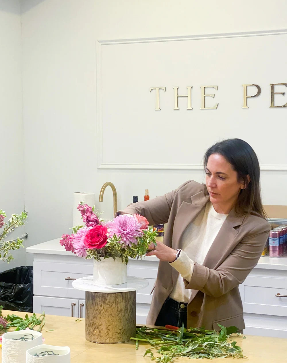 A professional florist in a tan blazer carefully styling a pink rose and chrysanthemum arrangement on a marble pedestal.