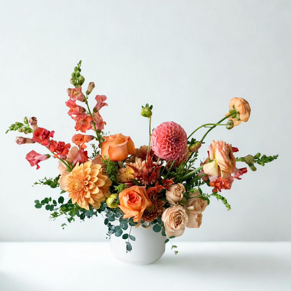 Signature autumn floral arrangement presented in a modern white, ceramic vase, showcasing a palette of bright coral, mauve, soft peach, and gentle orange blooms. Available for same-day delivery across Minneapolis and surrounding Twin Cities suburbs.